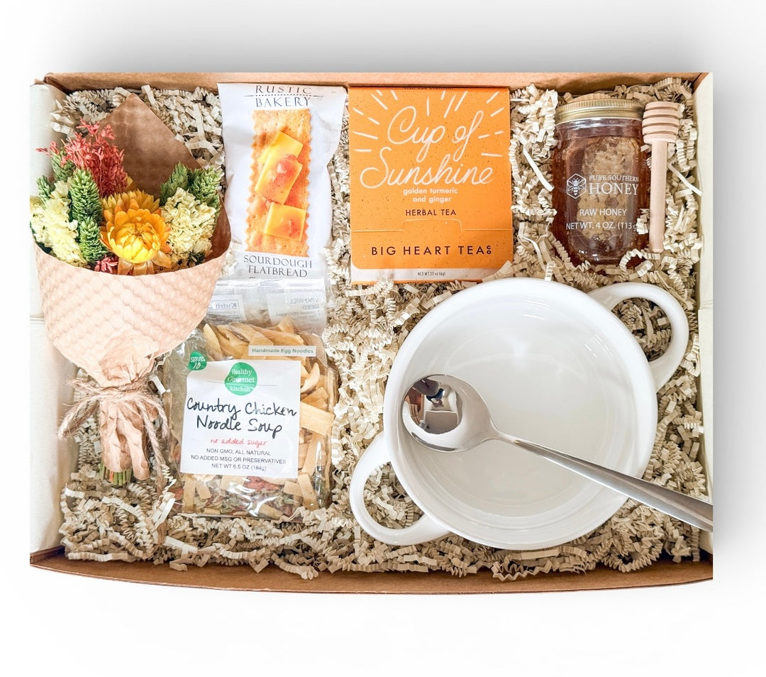 Curated gift box containing chicken noodle soup mix, sourdough flatbread, herbal tea, raw honey with dipper, white ceramic handled soup bowl with spoon, and a small dried floral bouquet arranged in crinkle paper.
