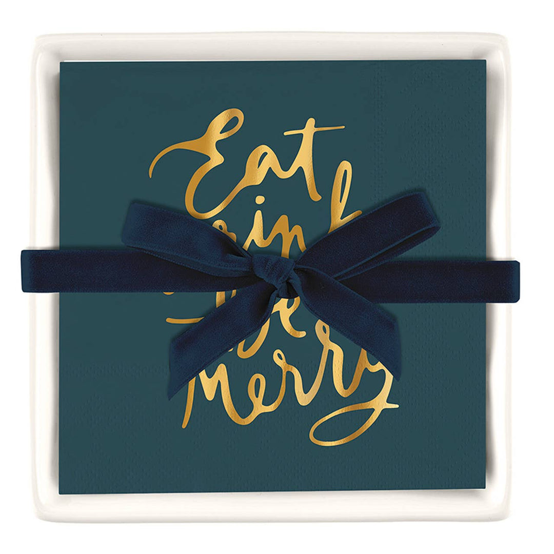 White napkin holder with teal napkins with gold text 'Eat, drink and be merry' and a navy ribbon.