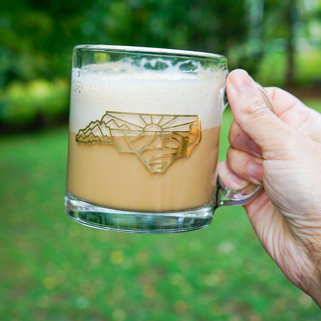 Glass mug with a geometric, mountain design of North Carolina held by a hand against a green outdoor background