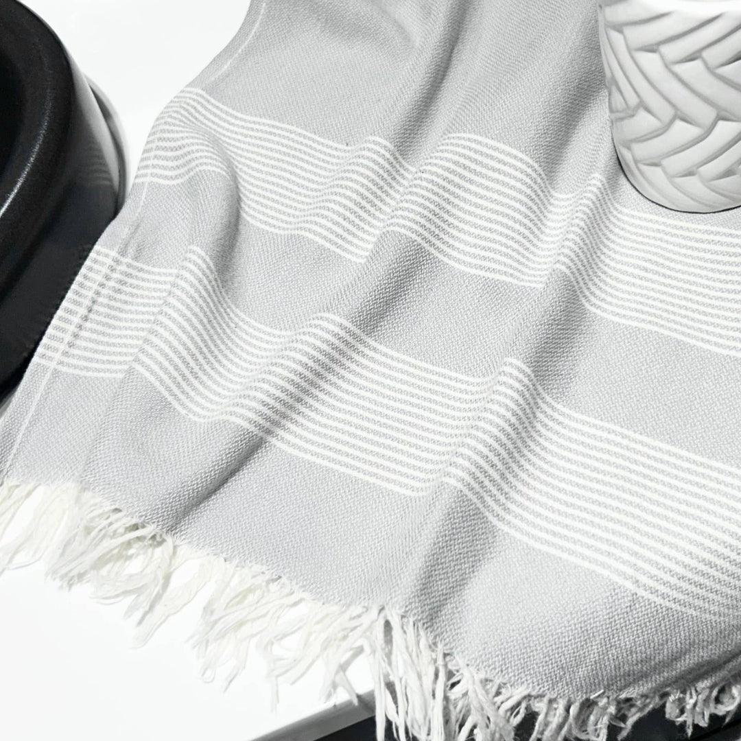 Grey towel with white stripes and fringe on a white surface. 