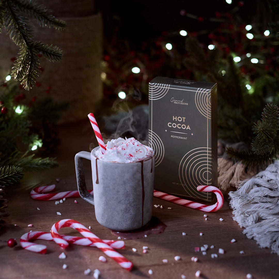 Hot chocolate with whipped cream and candy canes next to a Hot Cocoa box in a festive setting.