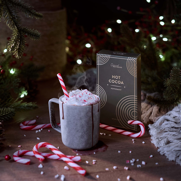 Hot chocolate with whipped cream and candy canes next to a Hot Cocoa box in a festive setting.