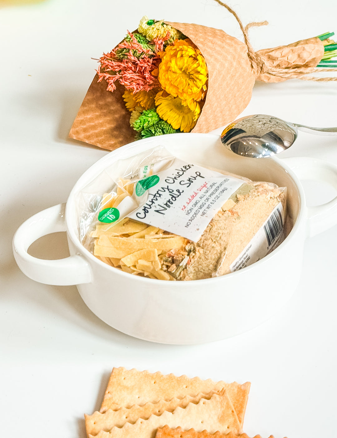 White ceramic handled soup bowl holding packaged chicken noodle soup mix with stainless steel spoon and a small dried floral bouquet in the background.
