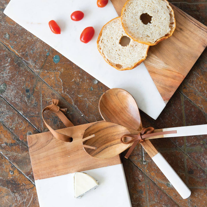 Wooden salad servers with a leather handle on a rustic wooden table with bread and tomatoes.