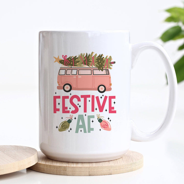 White mug with Christmas-themed design featuring a van and trees, placed on a white surface with a plant in the background.