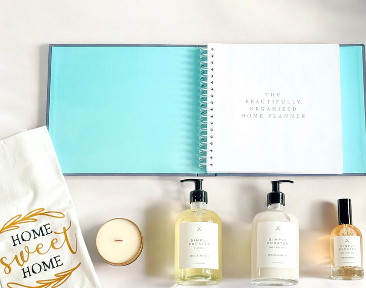 Flat lay of a luxury new home gift set with an open home organization planner, vanilla lavender hand wash, hand and body lotion, room and linen spray, soy candle, and a “Home Sweet Home” tea towel on a light background.