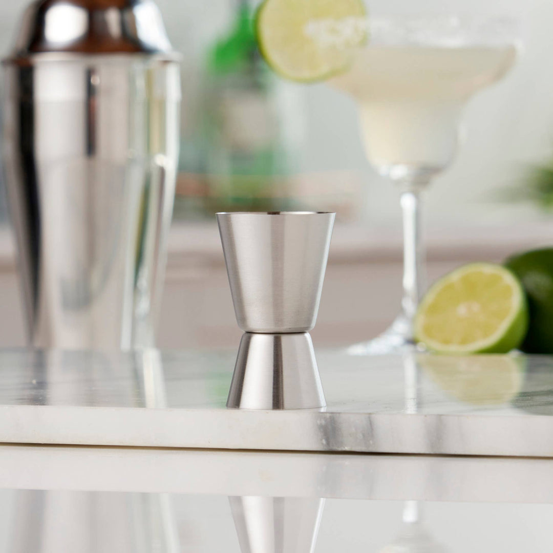 Metallic jigger on a bar counter with cocktail shaker, lime, and glass in the background