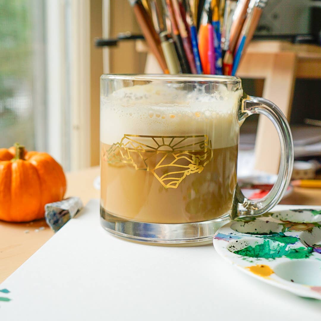 Clear mug with a geometric design of North Carolina filled with a creamy beverage, surrounded by art supplies on a table.
