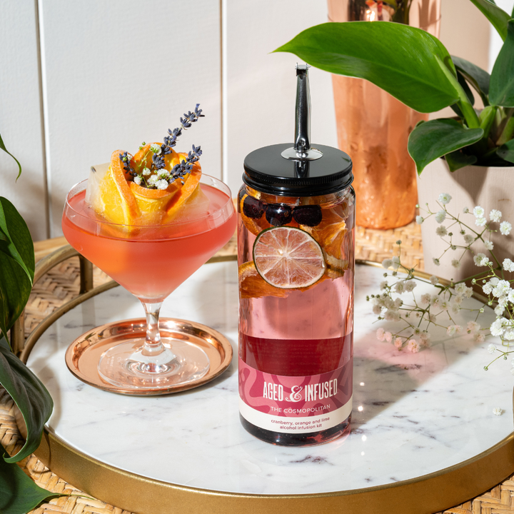 Cocktail in a glass with garnish next to a bottle of Aged &amp; Infused cocktail on a marble surface.