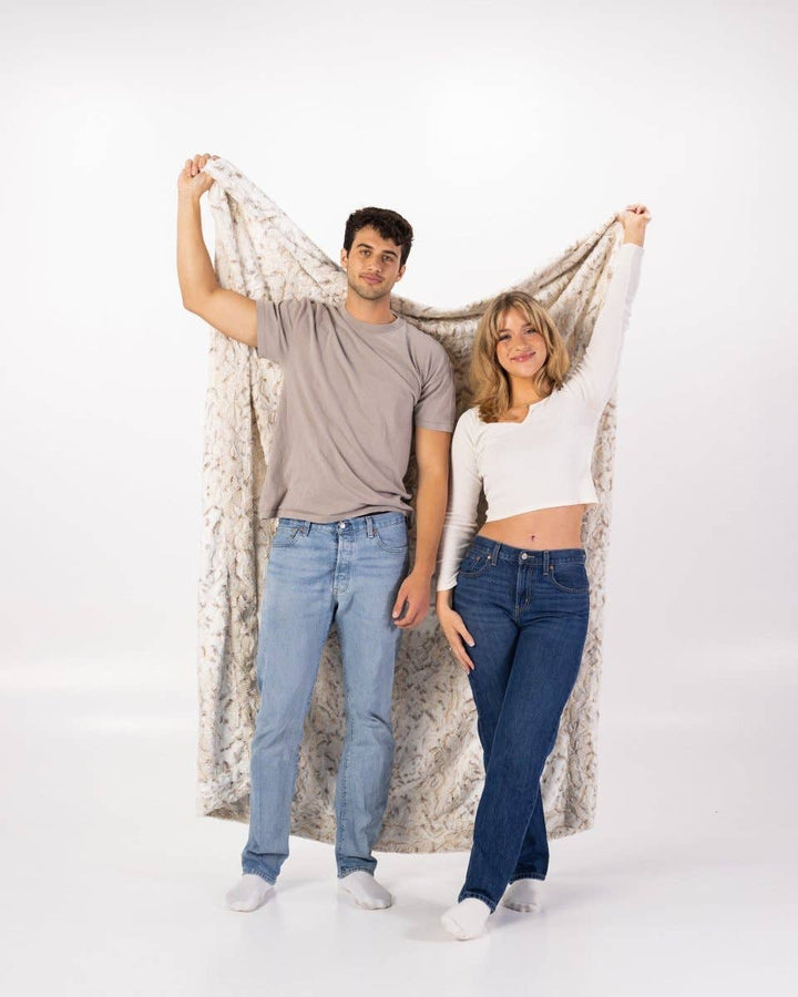 Two people standing in front of a white background holding a soft throw blanket. 