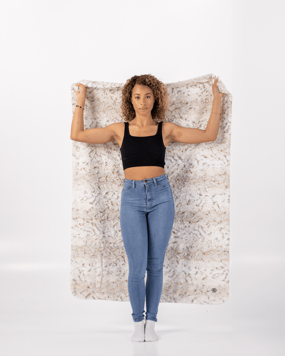 Woman wearing a black top and blue jeans standing against a white wall holding a soft throw blanket.