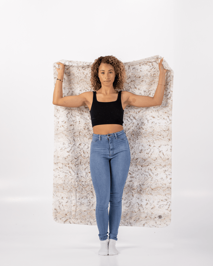 Woman wearing a black top and blue jeans standing against a white wall holding a soft throw blanket.