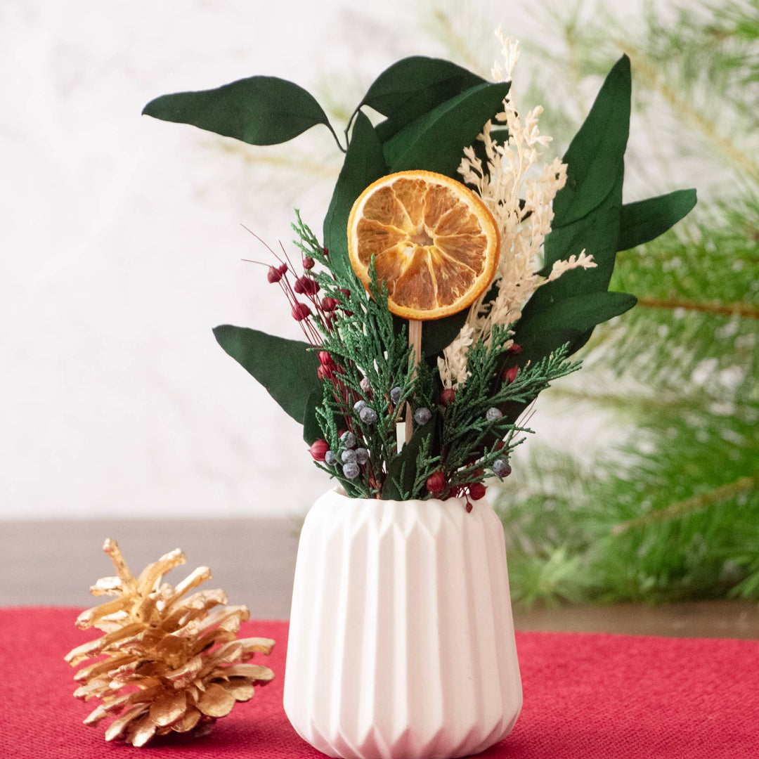 Decorative vase with dried flowers and an orange slice on a red surface with a blurred green background