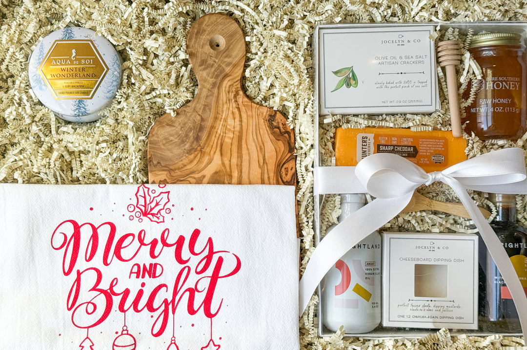 Gift basket featuring 'Merry and Bright' kitchen towel, 'Winter Wonderland' candle, wooden cutting board, olive oil and sea salt crackers, raw honey, sharp cheddar, oils, and a dipping dish. 