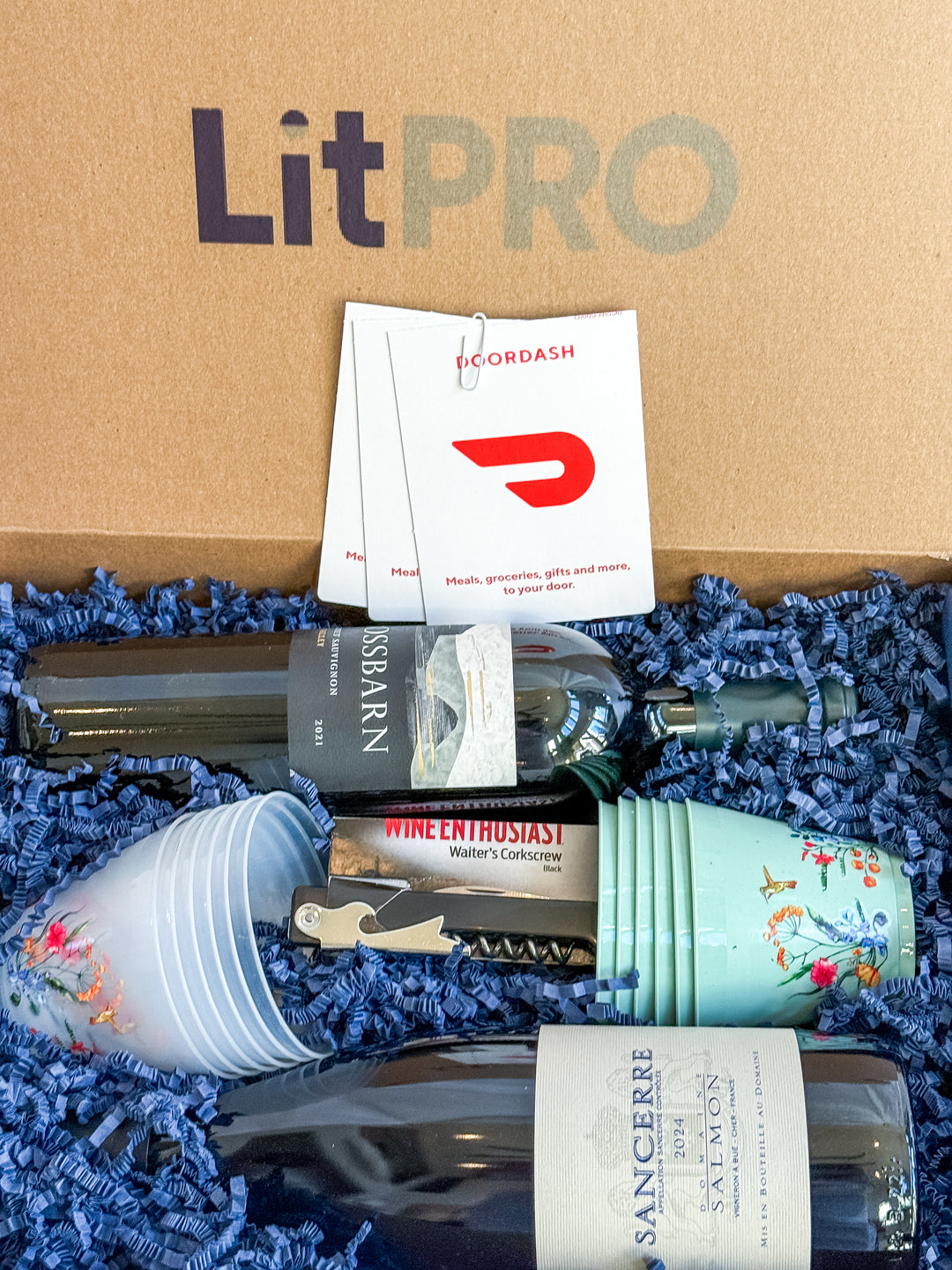 LitPro gift box with wine bottles, cups, a corkscrew and gift cards on blue crinkle paper. 