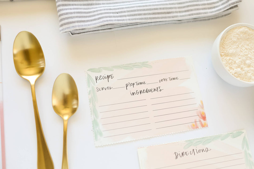 Recipe cards with gold spoons and a bowl of flour on a light surface