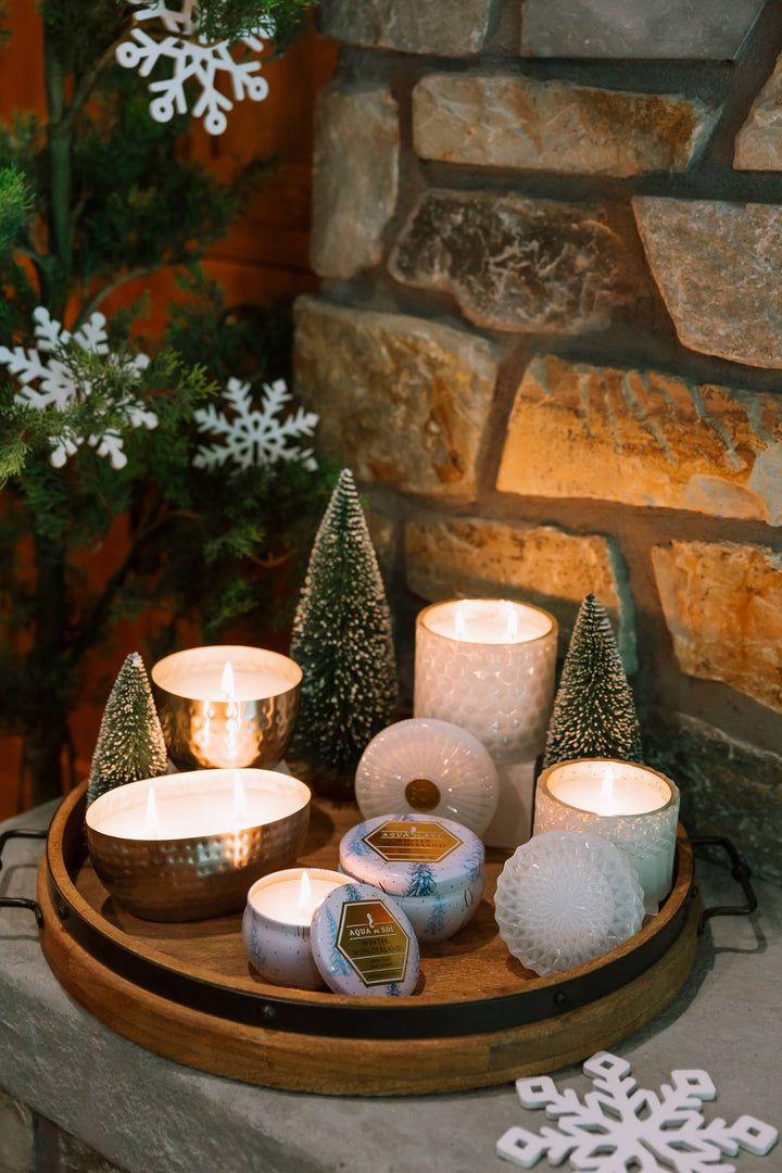 Decorative candles on a wooden tray with a stone wall and Christmas trees in the background.