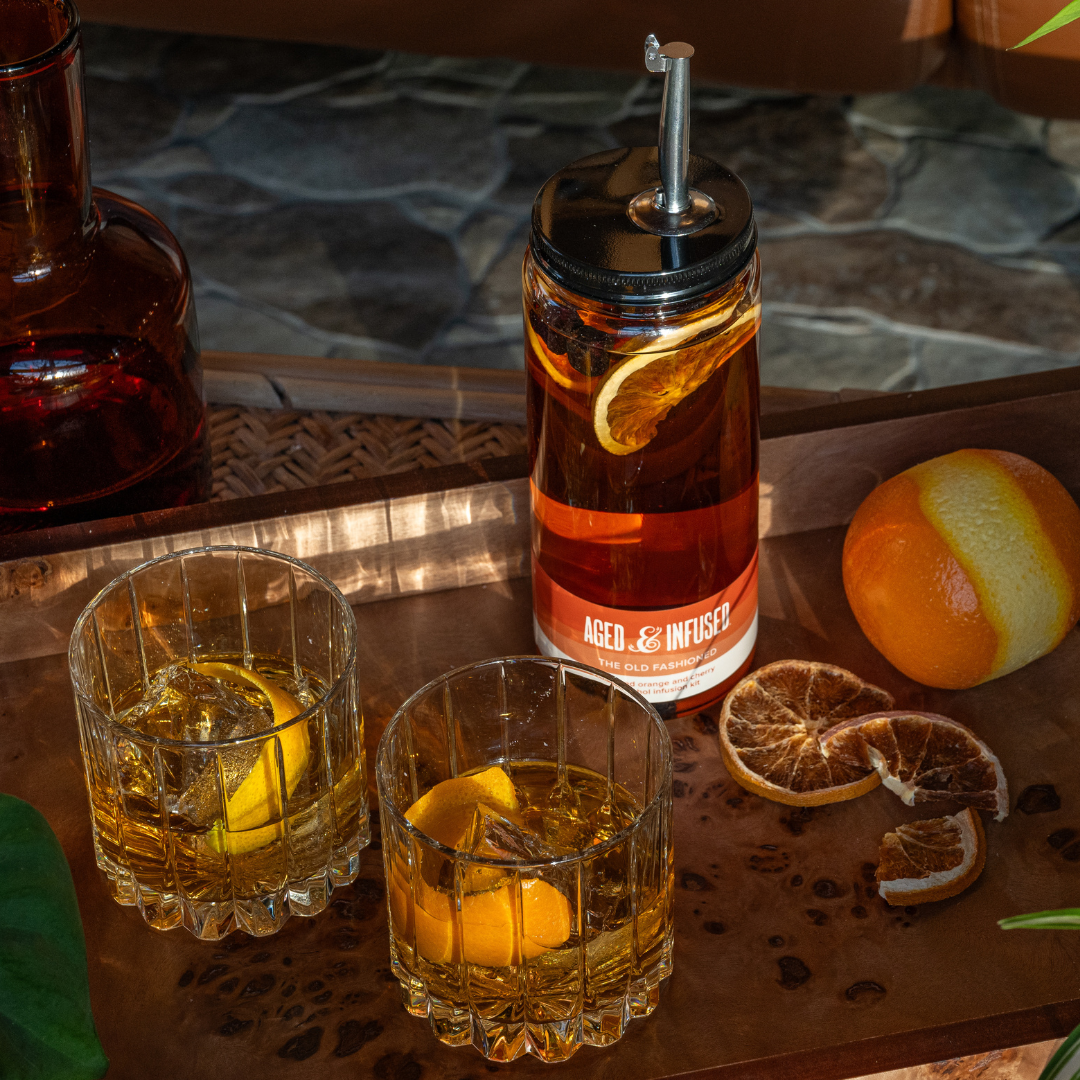 Bottle of 'Aged & Infused' whiskey with two glasses and oranges on a wooden surface.