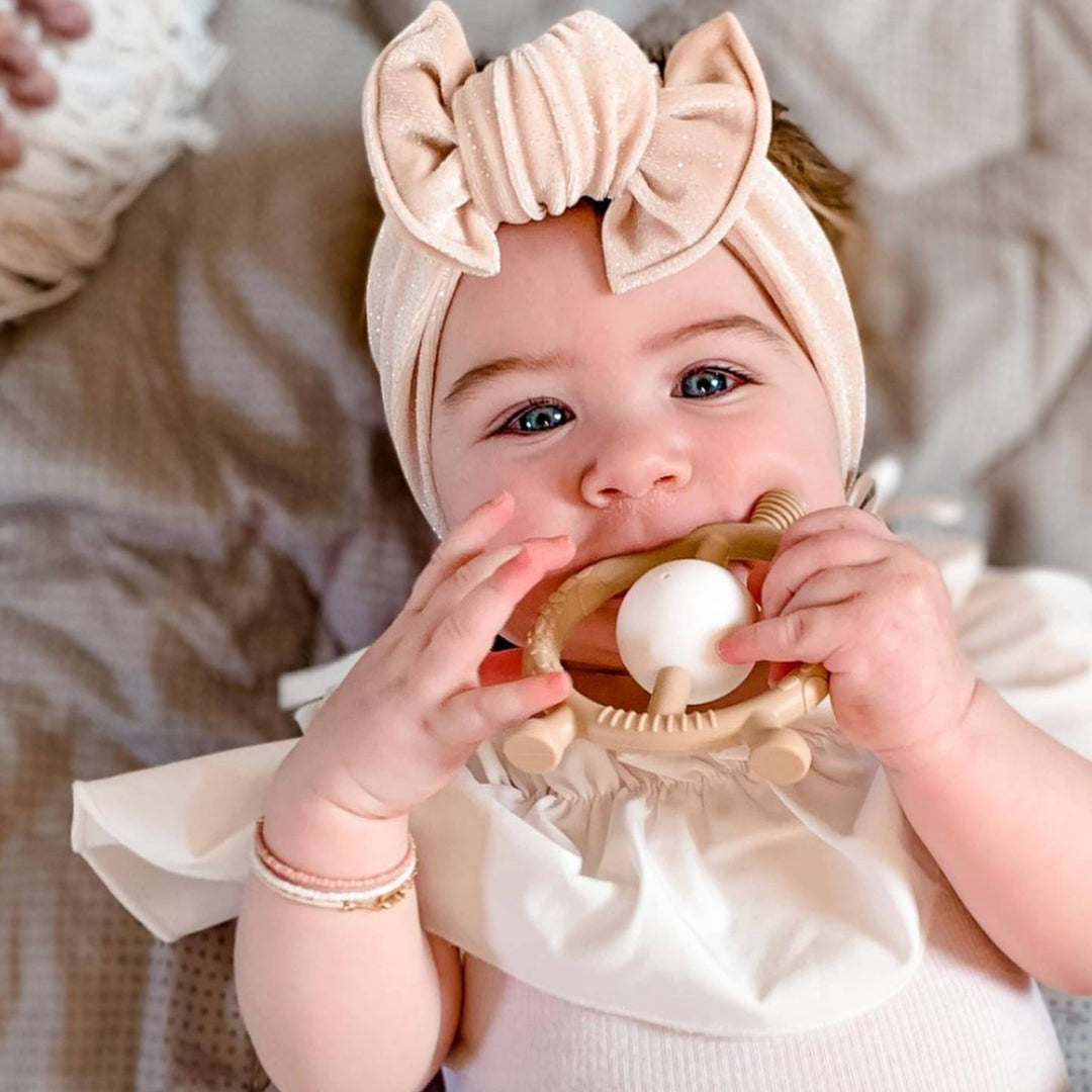 Baby wearing a beige headband with a bow, holding a teething ring.