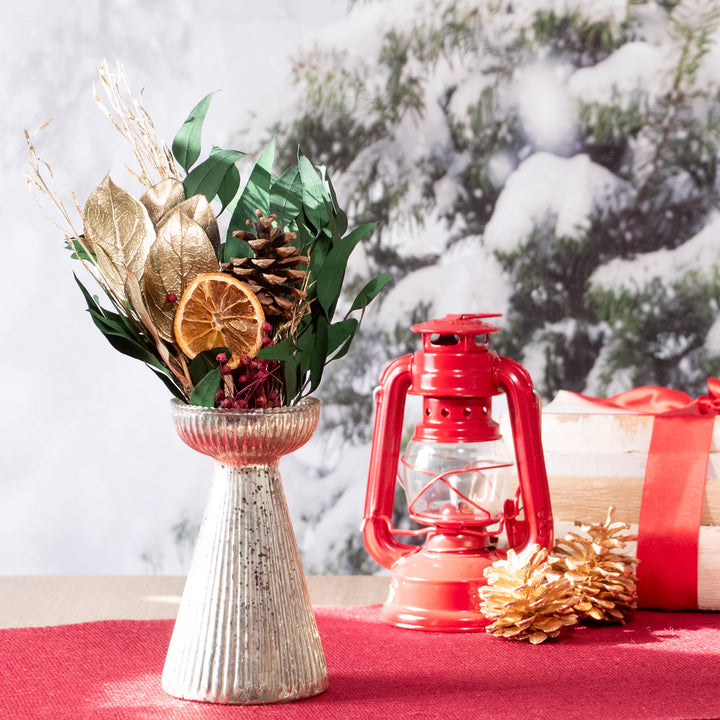 Decorative setup with a vase of dried leaves, a red lantern, and pinecones on a red surface with a snowy outdoor background.