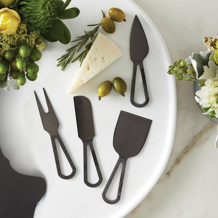 Set of black cheese knives on a white plate with cheese, olives, and herbs.