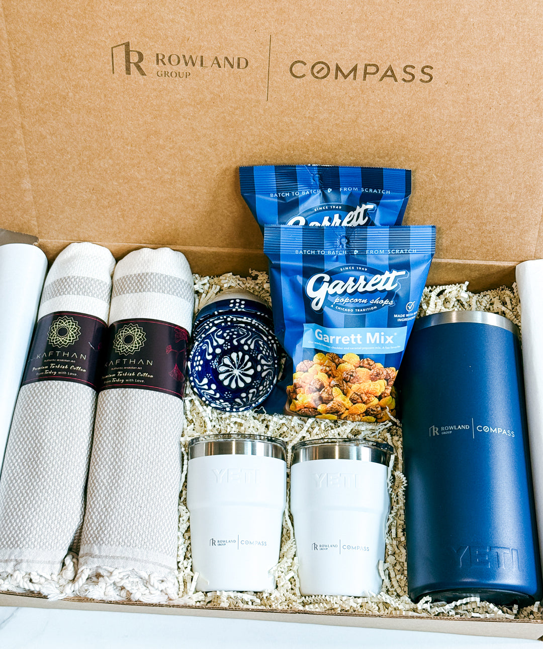 Gift set with Rowland Group and Compass branding, including towels, snacks, Yeti tumblers and French press.
