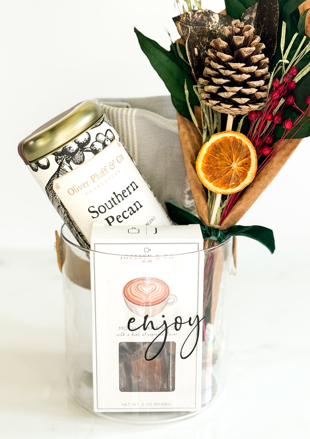 Glass container with coffee labeled 'Southern Pecan', a hand towel, mocha caramels and a bouquet of dried flowers on a white background