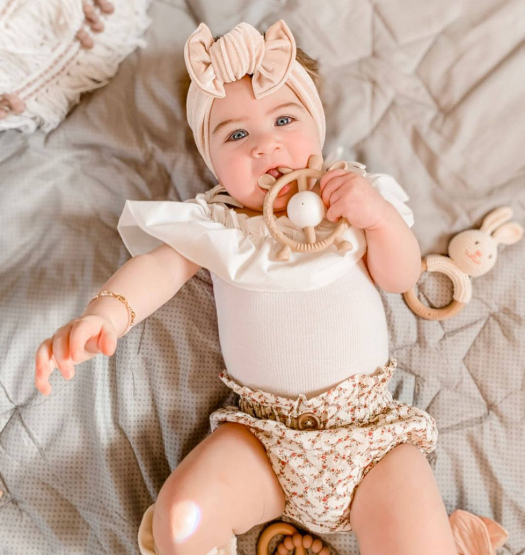 Baby lying on a textured surface wearing a white onesie with ruffles and floral diaper, holding a teething ring.