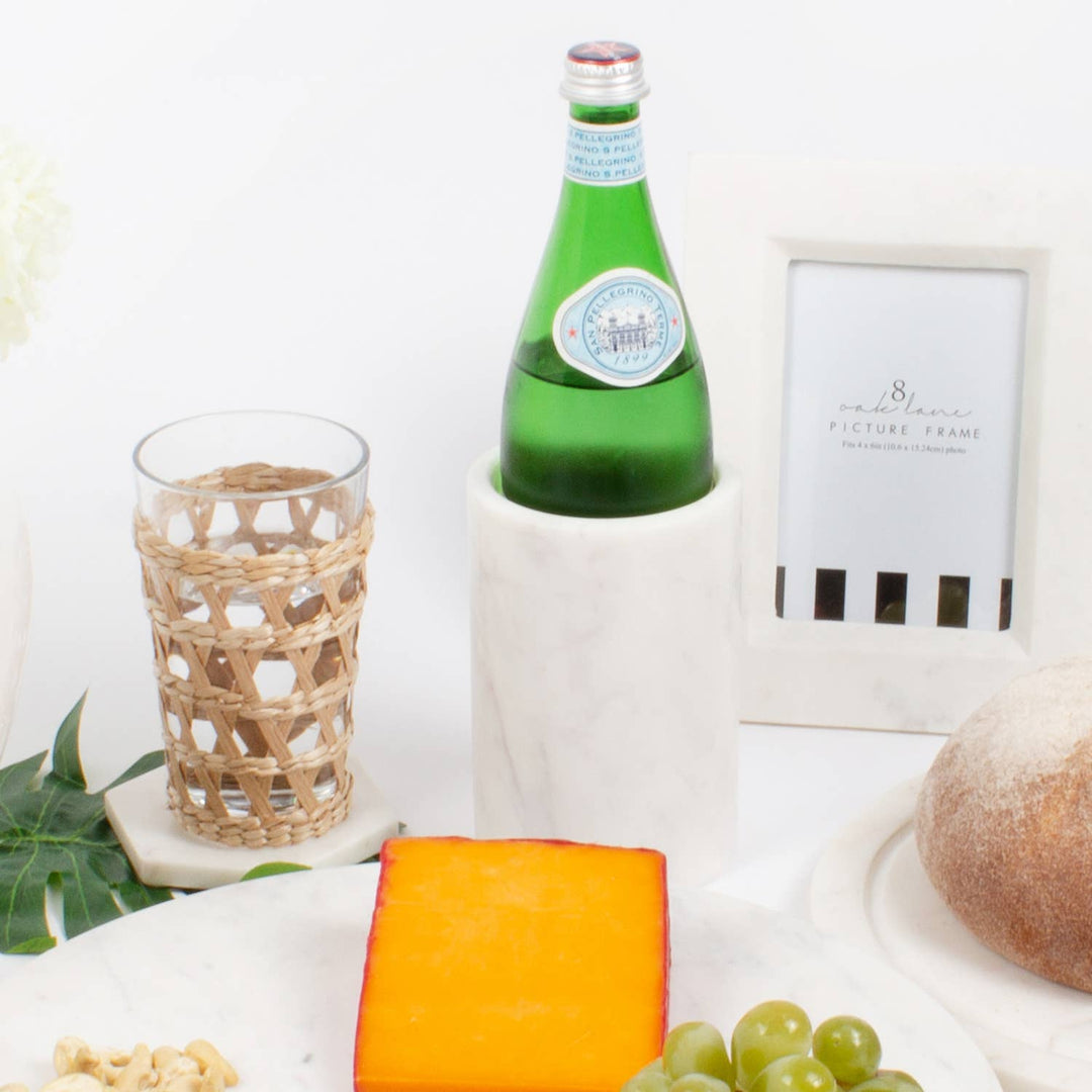 Green bottle in a marble cooler, glass, cheese, grapes, and bread on a white surface with a framed picture in the background.