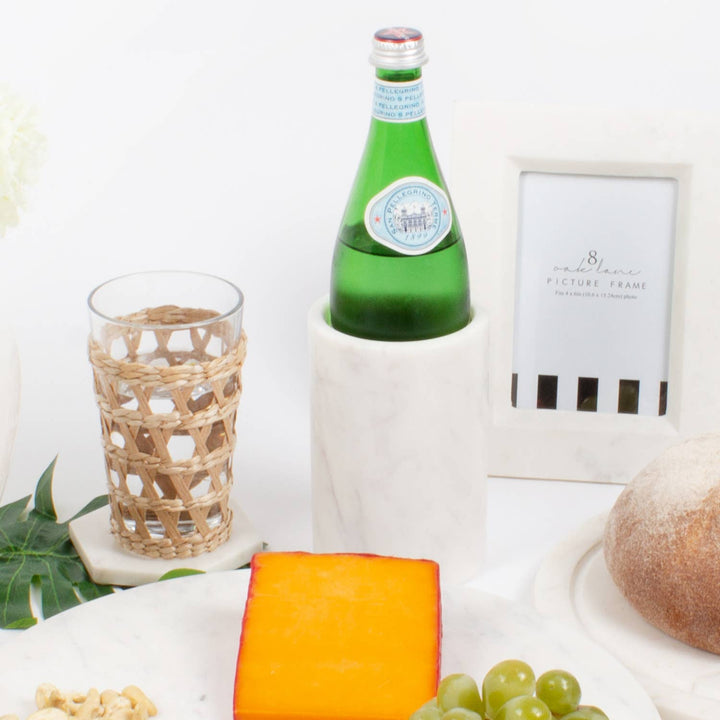 Green bottle in a marble cooler, glass, cheese, grapes, and bread on a white surface with a framed picture in the background.
