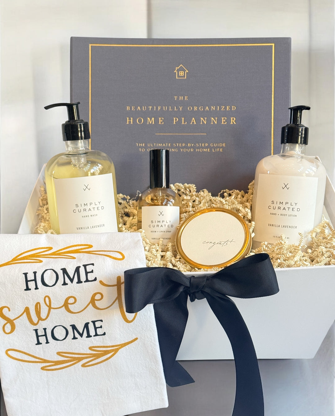 Luxury housewarming gift set in a white gift box with black ribbon, featuring The Beautifully Organized Home Planner, vanilla lavender hand wash, hand and body lotion, room and linen spray, soy candle, and a “Home Sweet Home” cotton tea towel.