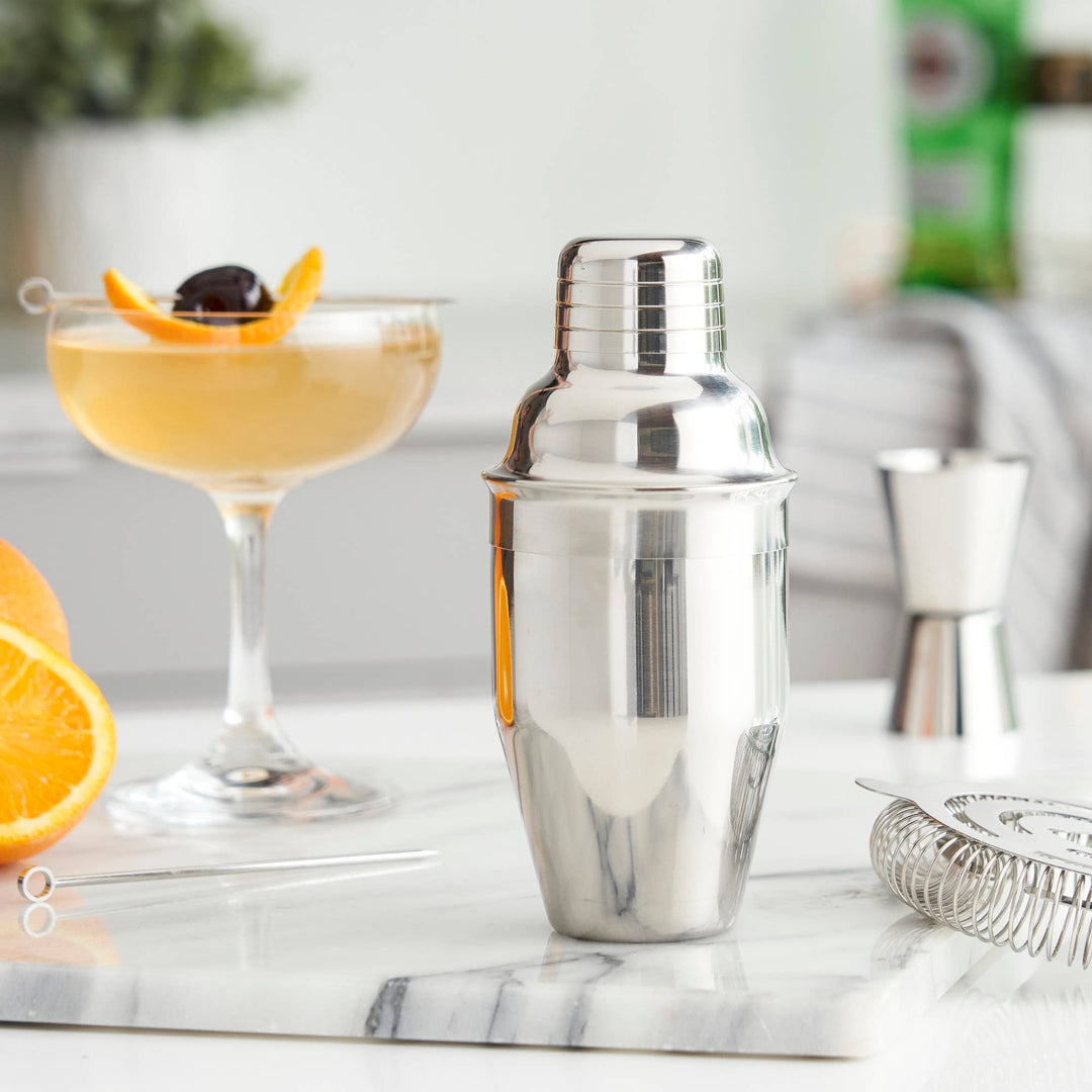 Silver cocktail shaker on a marble surface with a glass of orange cocktail and garnishes.