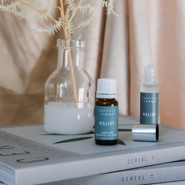 Two bottles of Thistle Farms Relief essential oil blend with a glass bottle and plant in the background.