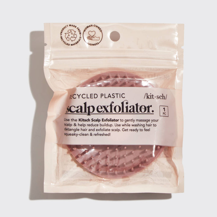 A terracotta-colored, plastic scalp exfoliator with flexible bristles in its plastic packaging with &