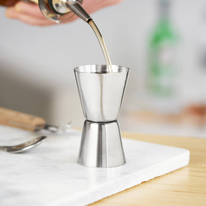 Metal jigger being filled with liquid on a marble surface