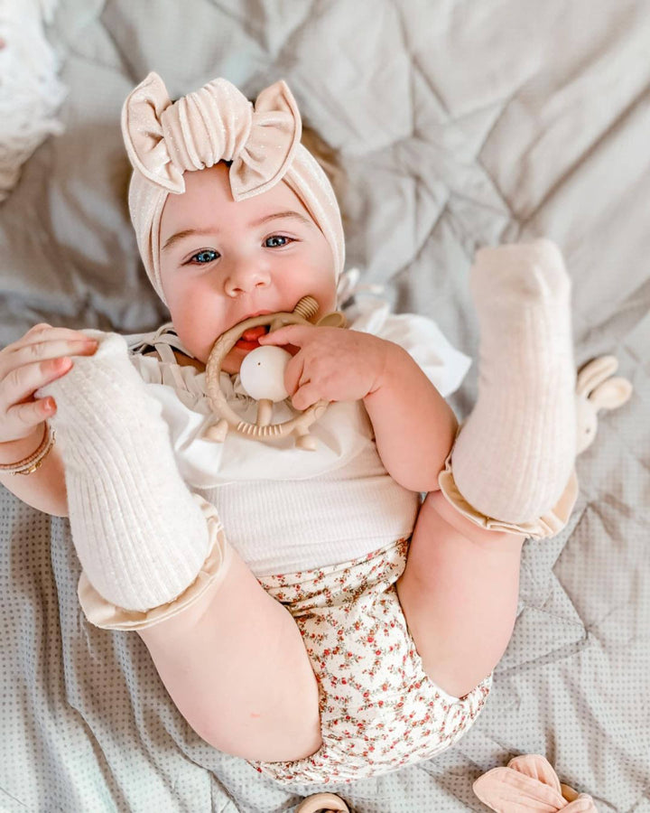 Baby lying on a soft surface wearing a white outfit with a bow and holding a teething ring.