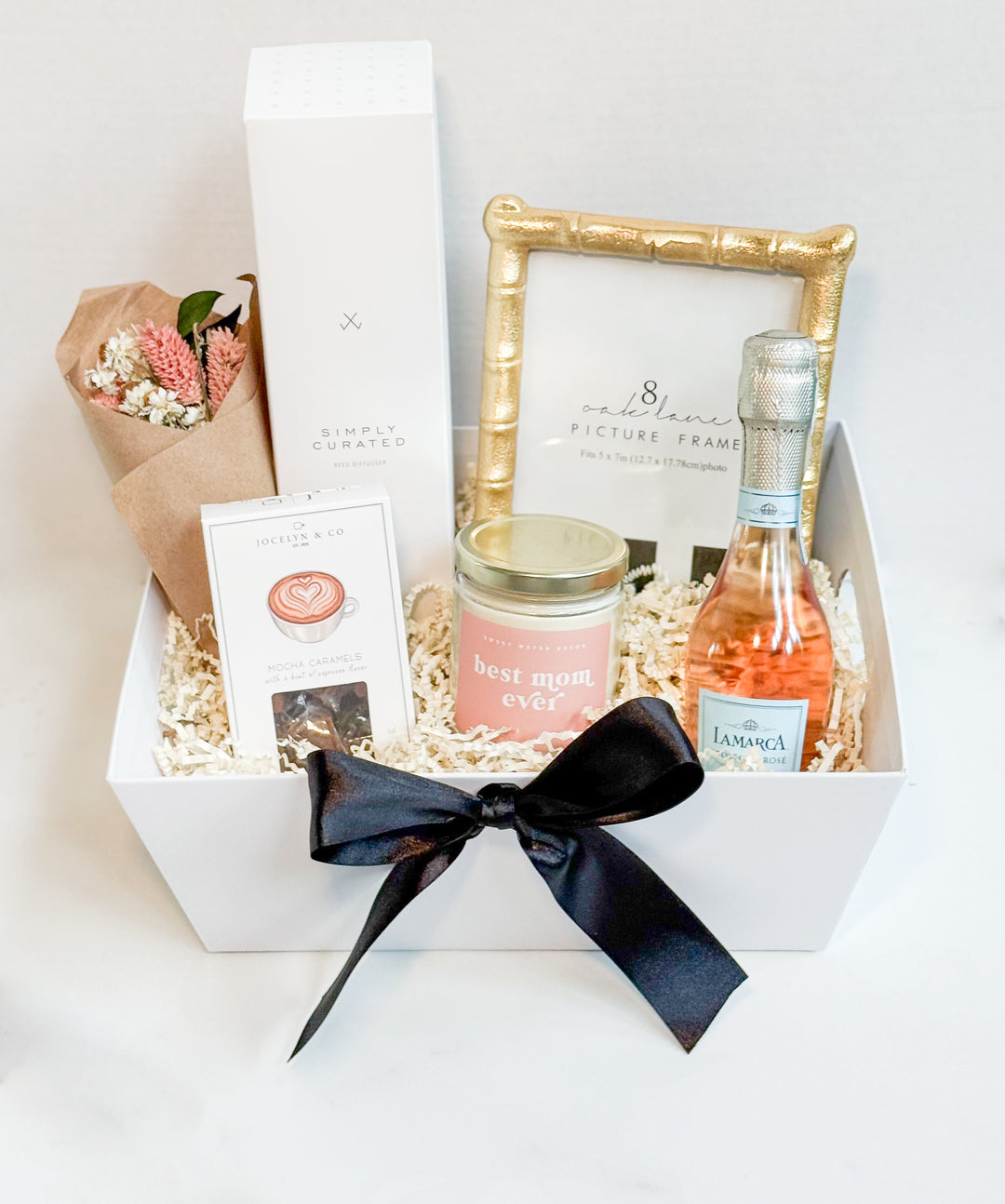 “Luxury Mother’s Day gift basket with soy candle labeled ‘Best Mom Ever’, mini pink dried bouquet, mocha caramels, vanilla lavender reed diffuser, gold picture frame, and mini bottle of La Marca Prosecco Rosé, all arranged in a white gift box with black ribbon – curated by Comfy-Cozy.
