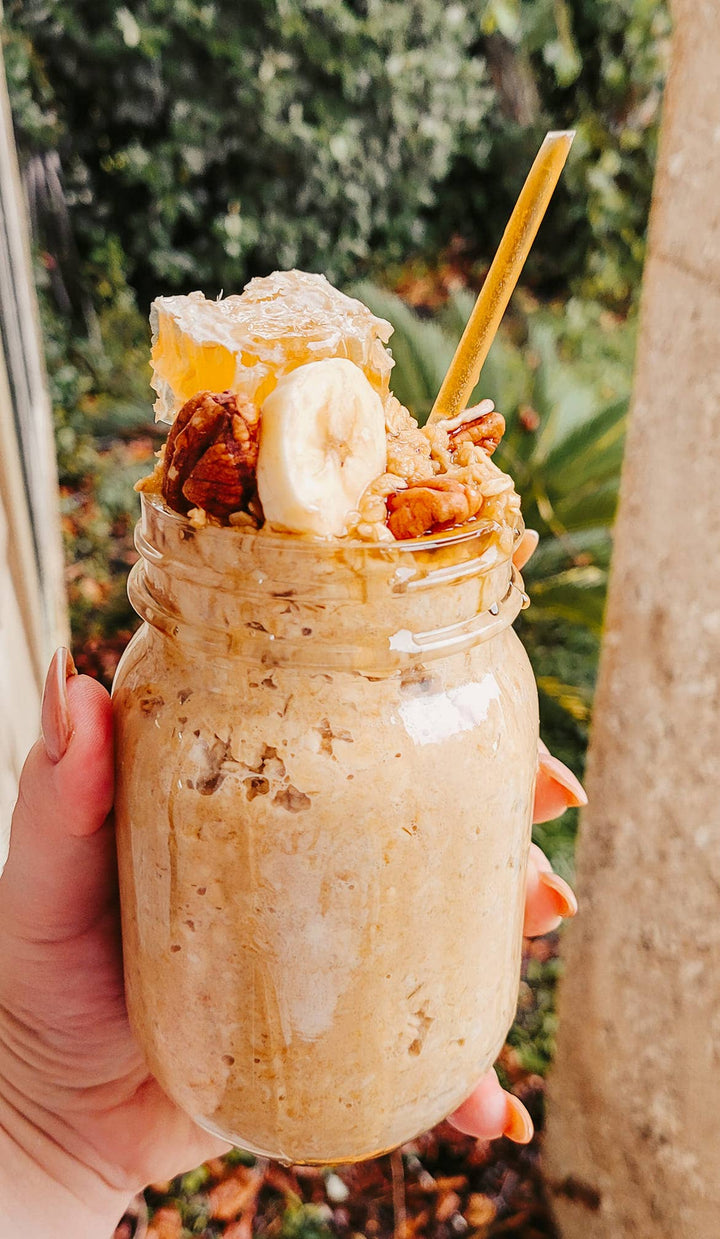 Mason jar with a layered dessert, including bananas, honey straw and nuts, held outdoors.