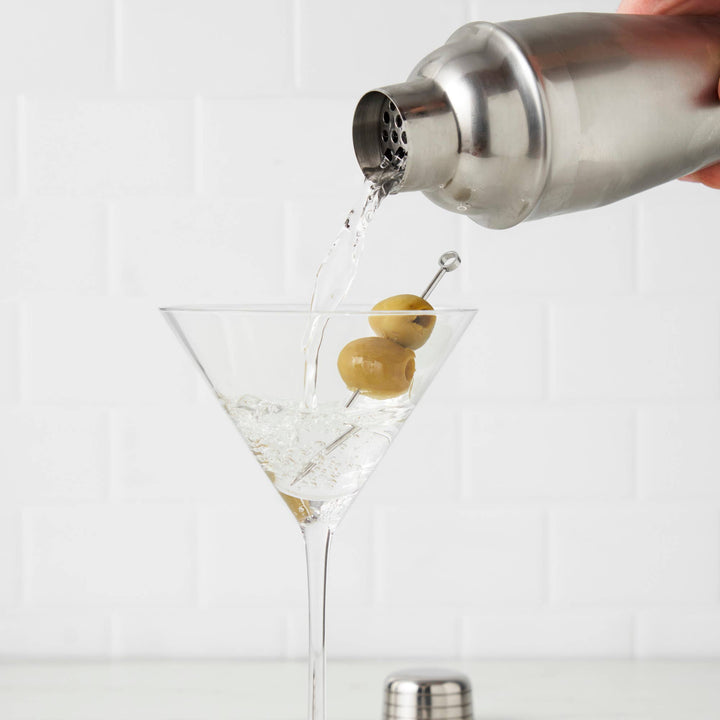 Cocktail being poured into a glass with olives using a shaker.