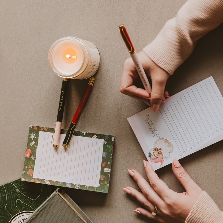 Person writing in a holiday-themed notebook with a lit candle and pens on a table