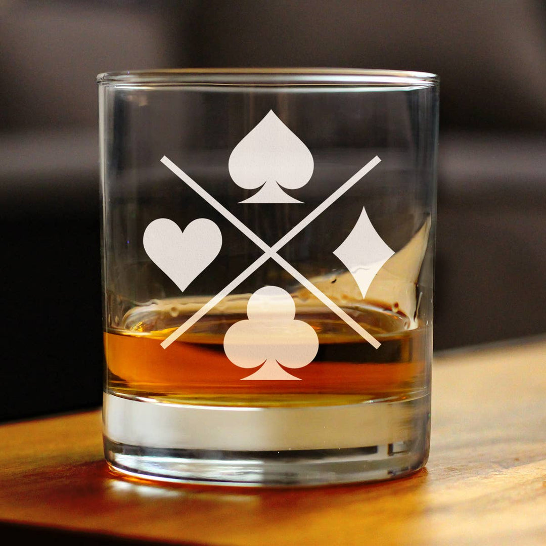 Clear whiskey rocks glass with playing card suits etched on it, filled with a drink, on a wooden surface.