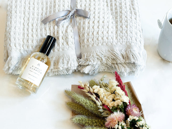 Room spray bottle, dried flowers, and a white textured blanket on a light surface