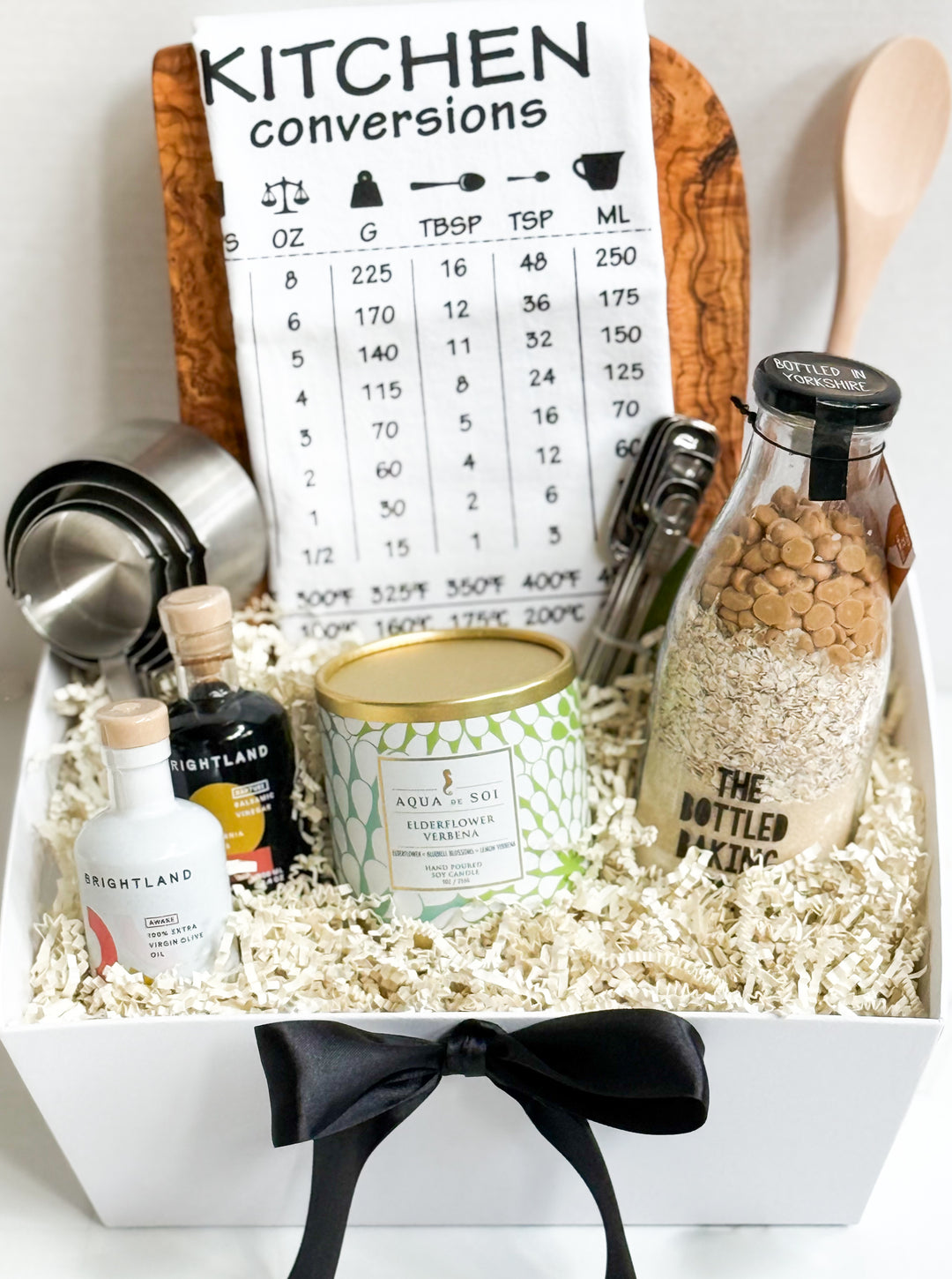 Gift basket with cutting board, cookie mix, and kitchen items including a bottle of oil, a canister, and a measuring cup on a white background.