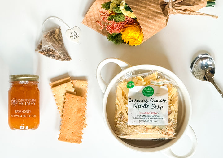 Top view of chicken noodle soup mix in white ceramic bowl surrounded by sourdough flatbread, raw honey jar, herbal tea bag, stainless steel spoon, and dried floral bouquet on a neutral surface.