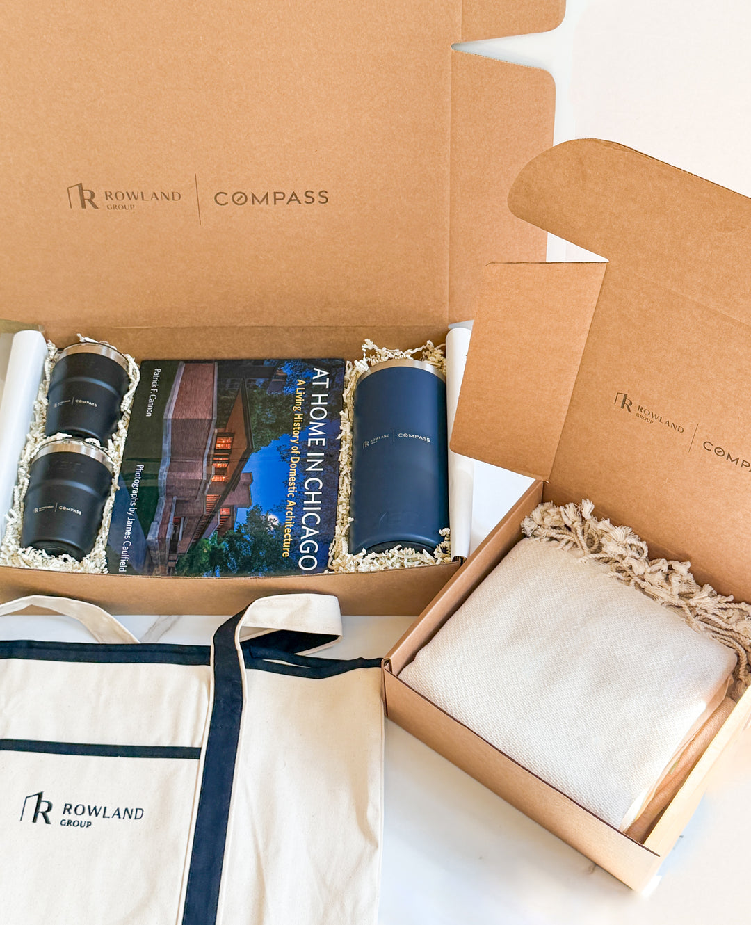 Open box containing Rowland Compass products including a book, a coffee press, tumblers, a blanket and a tote bag.