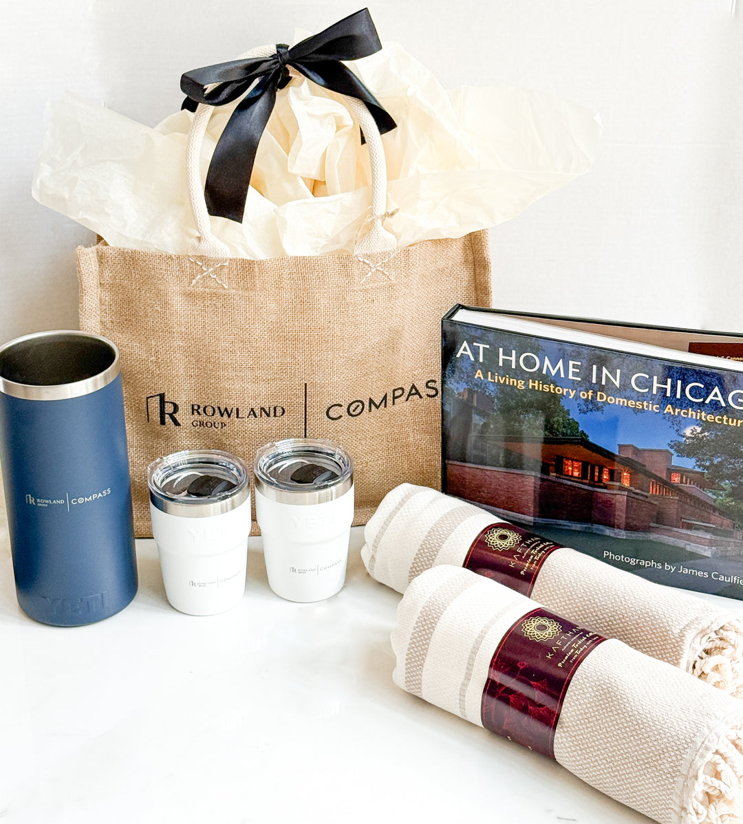 Gift bag with Rowland Compass branding, tumblers, a book, and towels on a white surface.