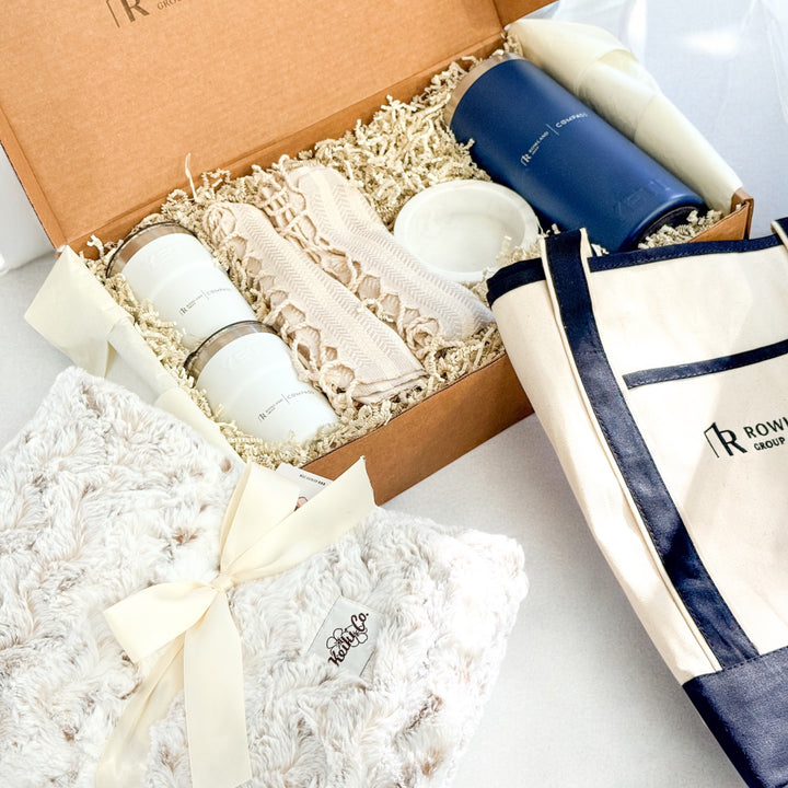 A "Rowland Group" gift set with 2 white tumbler cups, a blue large tumbler, a beige blanket with fringe and a tote bag on a white surface.