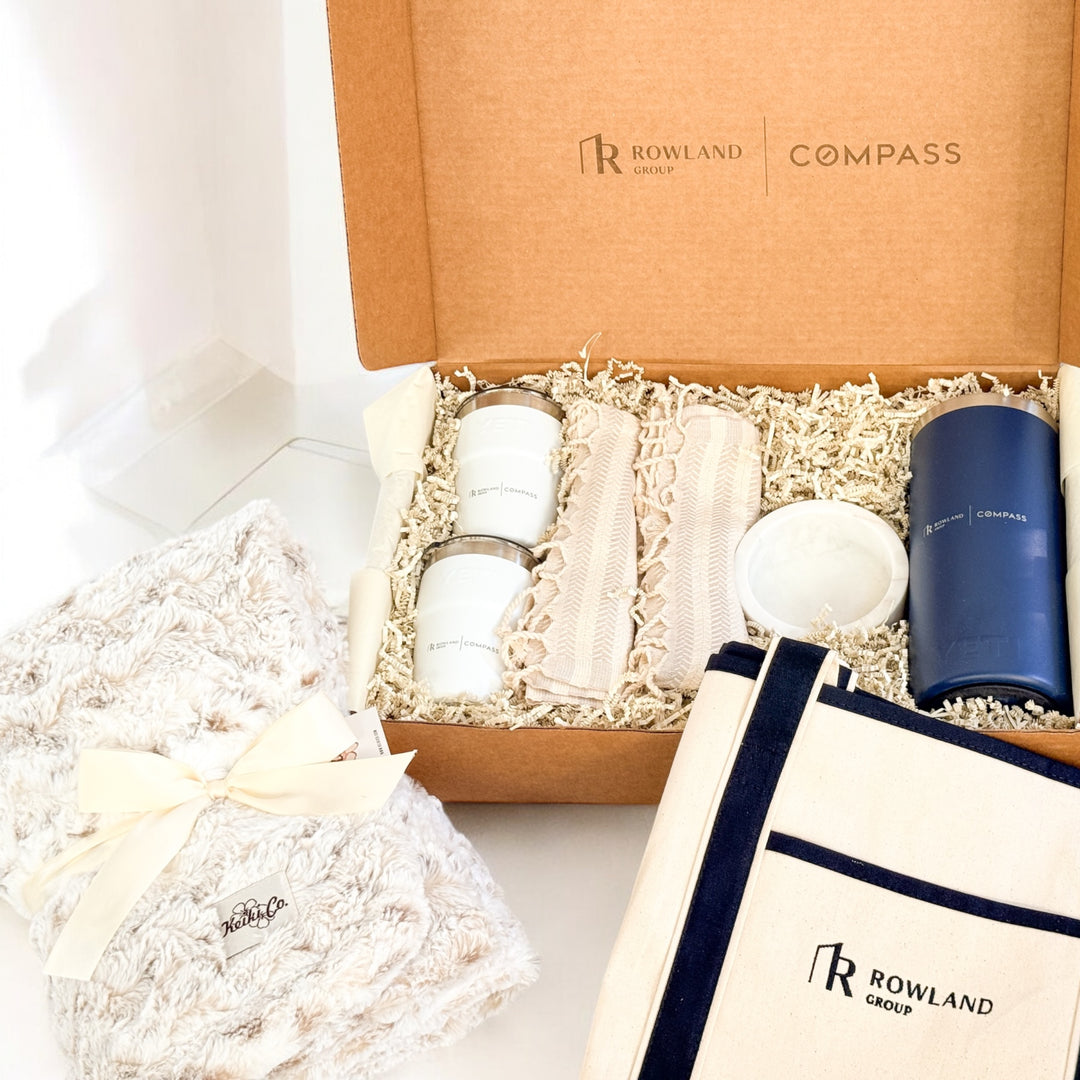 Gift set including a blue YETI tumbler, white tumbler cups, a beige blanket with fringe and a white "Rowland Group" tote bag on a light background.