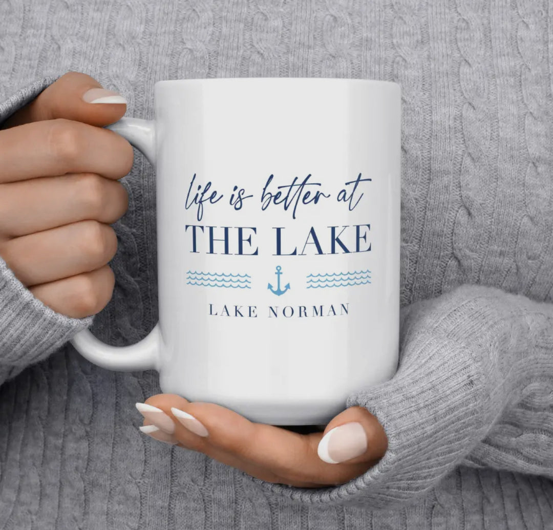 White mug with 'life is better at THE LAKE' text held by a person wearing a gray sweater.