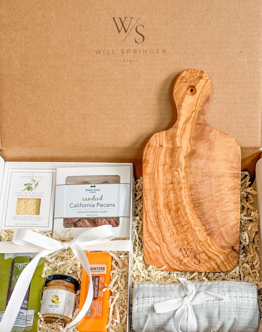 Gift box with wooden cutting board, snacks, and a grey towel in a brown box.